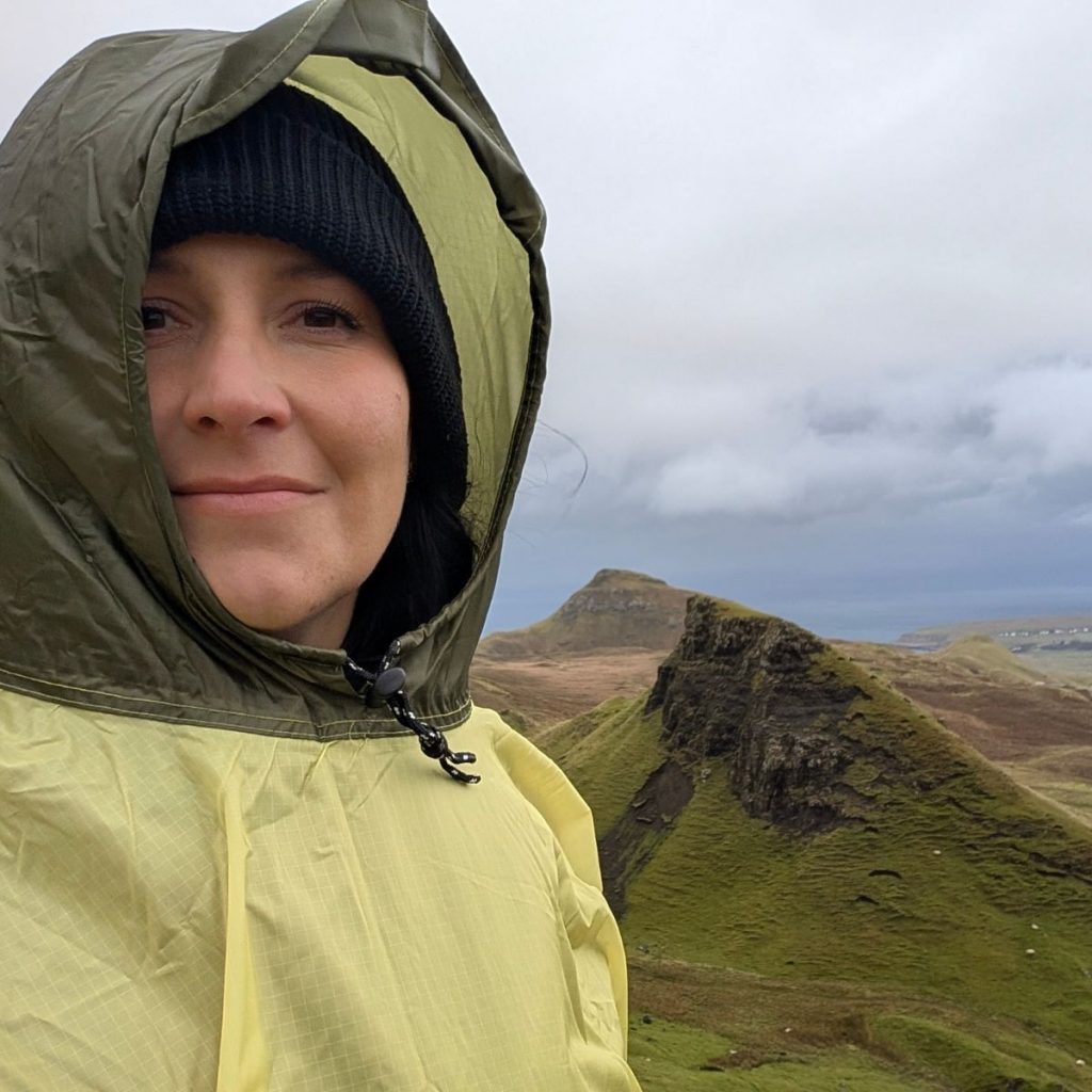 Frau mit schwarzer Mütze und gelbem Regencape steht im Regen vor grünen Bergen – Symbol für authentisches Marketing statt Perfektion.