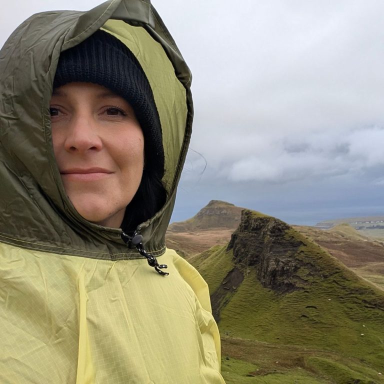 Frau mit schwarzer Mütze und gelbem Regencape steht im Regen vor grünen Bergen – Symbol für authentisches Marketing statt Perfektion.