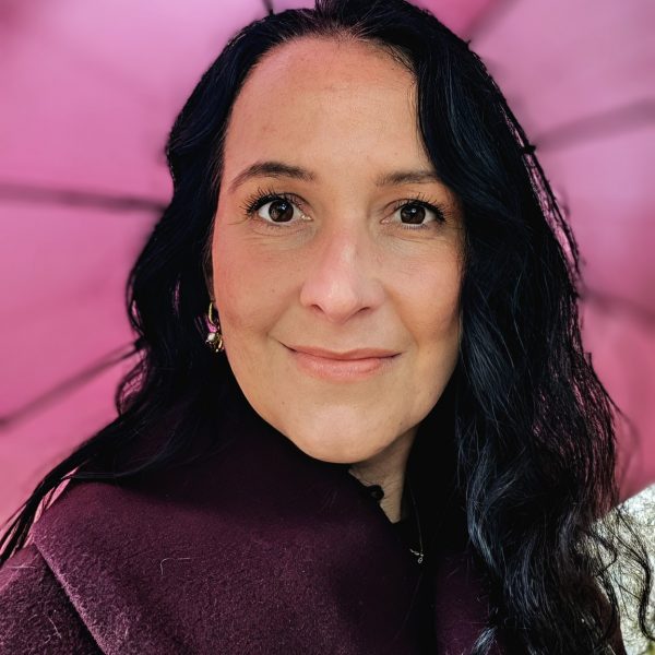 Anne Hofmann trägt einen lilafarbenen Mantel und hält einen passenden Schirm. Sie lächelt in die Kamera, während der Wind durch die schottische Landschaft zieht.